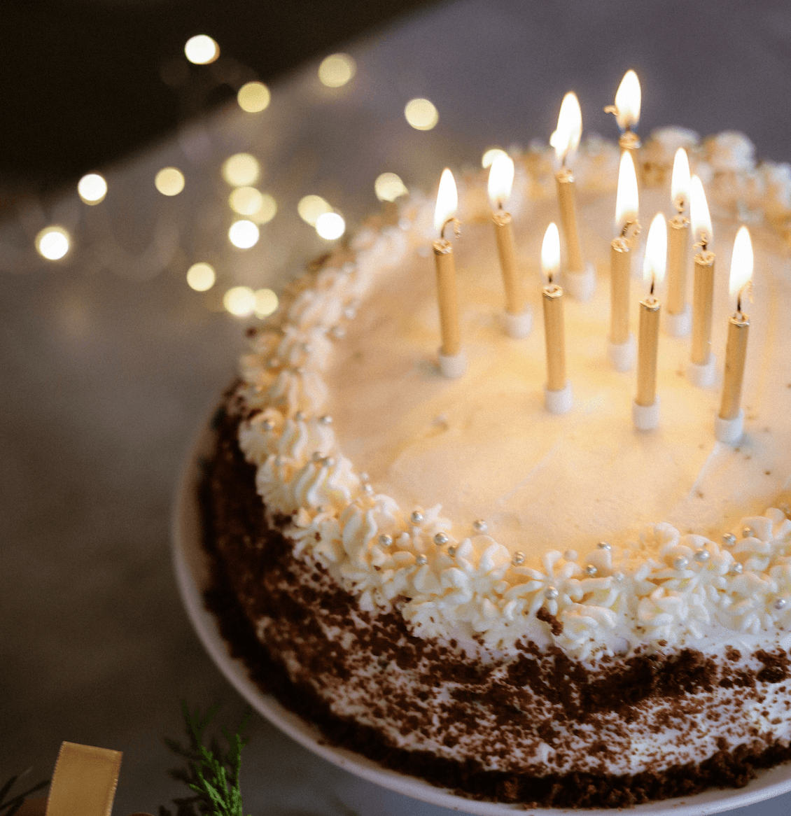 Torta sa šlagom, srebrnim jestivim perlama i upaljenim svećicama, ispred zamagljenih svetlucavih lampica
